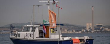 Barco de comité 'Willie Piccone' del Royal Gibraltar Yacht Club (RGYC) fondeado en la Bahía de Algeciras, mostrando banderas de la regata y el logotipo 'Interclubs del Estrecho'. De fondo se aprecian las grúas del puerto y la central térmica, con la costa difuminada por la bruma.